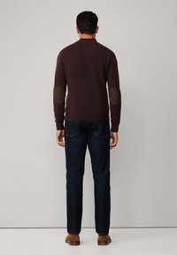 Maglione marrone scuro con toppe ai gomiti, polsini a coste e design attillato, abbinato a jeans blu scuro e scarpe marroni.