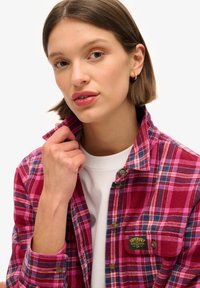 Chemise en flanelle à carreaux rouges et roses avec un design à boutons, comportant une poche poitrine et un petit patch de marque. Portée par-dessus une chemise blanche.