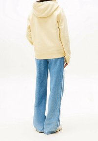 Keltainen huppari, joka on yhdistetty leveälahkeisiin sinisiin farkkuihin, joissa on valkoinen raita sivuilla. Rentoa tyyliä.