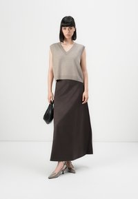 Woman with straight dark hair wearing a beige sleeveless V-neck sweater, long dark skirt, gray heels, holding a black handbag against white background.