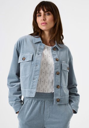 Frau in einer hellblauen Cord-Jacke und passenden Hosen über einem weißen gemusterten Oberteil, die mit den Händen in den Taschen vor einem weißen Hintergrund steht.