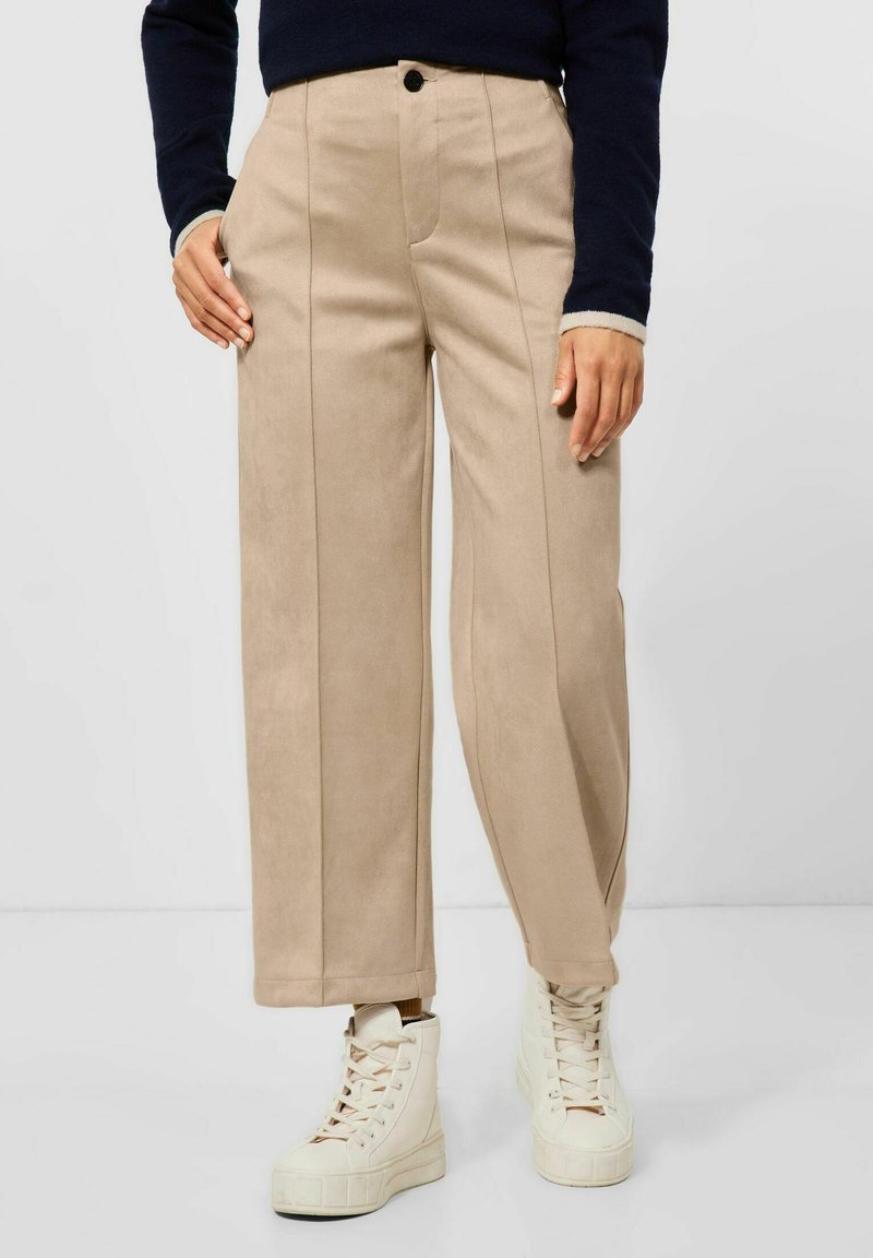 Pantaloni beige a gamba larga realizzati in materiale morbido, con dettagli di cuciture verticali e chiusura frontale con bottone, abbinati a sneakers alte bianche.