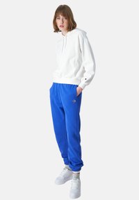 Felpa bianca con cappuccio, pantaloni sportivi blu con bordo elastico e sneakers bianche. L'abbigliamento presenta dettagli con logo e tasche laterali.