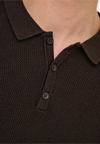 Brązowa polo z kołnierzykiem, dwoma czarnymi guzikami i wzorem tkanym. Tkanina wydaje się miękka i elastyczna.