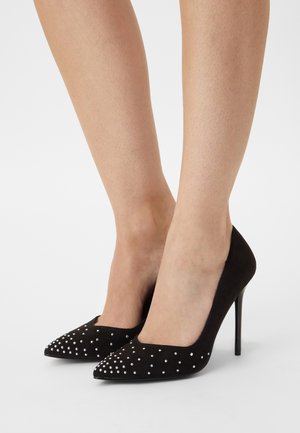 Talons aiguilles en suede noir avec des bouts pointus, ornés de petits clous en argent à l'avant, portés sur des jambes nues sur fond blanc.