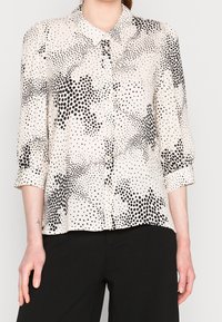 Blusa beige chiaro con un motivo a pois neri, dotata di chiusura con bottoni, colletto e maniche arrotolate. Tessuto liscio.