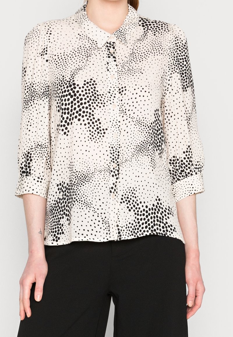 Blusa beige chiaro con un motivo a pois neri, dotata di chiusura con bottoni, colletto e maniche arrotolate. Tessuto liscio.