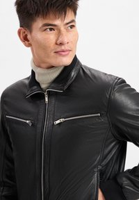 Zwarte leren jas met een gladde textuur, voorzien van een korte kraag, zilveren ritszakken en geribde manchetaccenten.