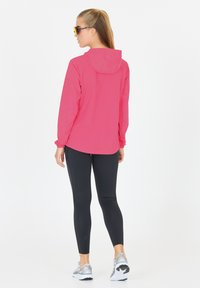 Lätt, rosa jacka med huva och långa ärmar, kombinerad med svarta leggings med texturerade detaljer och grå träningsskor.