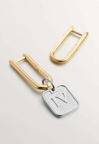 Un hardware en forma de clip de papel en color dorado y un charm rectangular plateado con una letra "N" en relieve cuelgan juntos.
