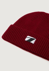 Gorro de punto rojo con textura acanalada, que presenta una etiqueta blanca con un acento gráfico en blanco y negro.