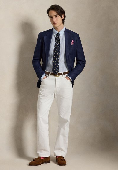 Noor mees kannab tumesinist jopet, triibulist särki, mustriga lipsu, valgeid pükse, pruune tutiga mokasiine ja roosat taskurätti, seisab käed taskutes.