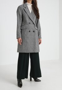 Doppelt brechender Mantel mit einem schwarz-weißen Hahnentrittmuster, ausgestattet mit einem Reverskragen, kombiniert mit weit geschnittenen, dunkelgrünen Hosen.