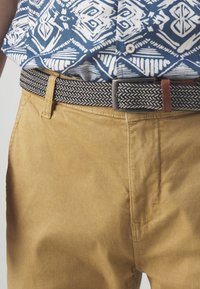 Pantaloni din bumbac bej deschis cu o textură netedă, asortați cu o curea țesută navy cu un model, având accente gri și ruginiu.