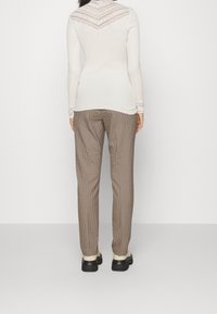 Vrouw in een crèmekleurig topje met kant, lange mouwen, bruin geruite broek en crèmekleurige schoenen met zwarte zolen, staande met de rug naar de camera op een witte achtergrond.