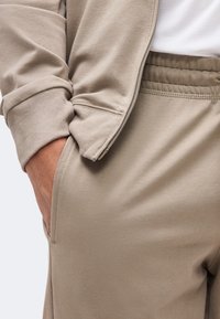 Άτομο που φοράει μπεζ φόρμες και τζάκετ με φερμουάρ που ταιριάζει, με το χέρι στην τσέπη, δείχνοντας την υφή του υφάσματος και τις λεπτομέρειες των ραφών.