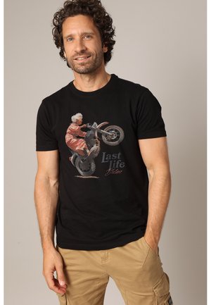 Man with curly hair wearing black t-shirt with graphic of elderly person doing a motorcycle wheelie and text "Last Life Deluxe".