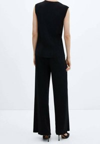 Žena stojící zády, oblečená v černém tílku bez rukávů, širokých černých kalhotách a černých vysokých podpatcích na jednobarevném bílém pozadí.