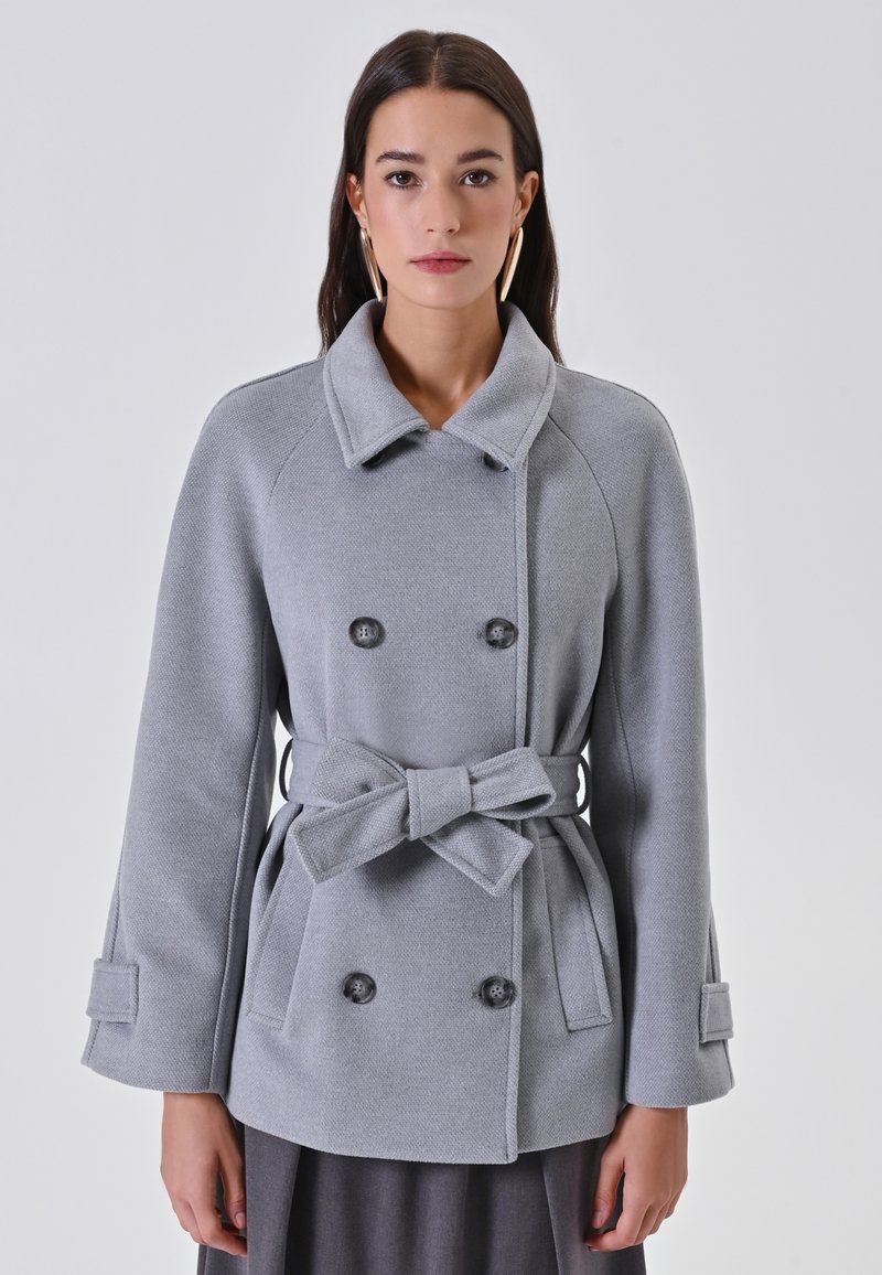 Cappotto grigio a doppio petto con cintura legata, ampio colletto e due grandi bottoni; realizzato in tessuto testurizzato con tasche laterali e maniche lunghe.