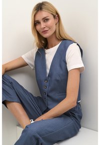 Blå chambray väst med djup V-urklippning, fem bruna knappar och två sidofickor, kombinerad med en vit t-shirt och löst sittande byxor.