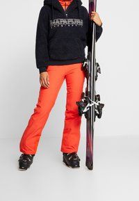 Blaue Fleece-Hoodie mit Logo, leuchtend orangefarbene Skihosen und schwarze Skistiefel. Skier mit lila Akzenten in der rechten Hand.