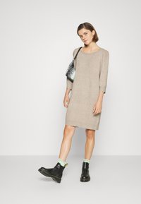 Abito in maglia beige con maniche a tre quarti e vestibilità rilassata. Il modello indossa stivaletti neri alla caviglia con calzini in pizzo verde menta e una borsa a fantasia.