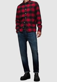 Camicia di flanella a quadri rossa e nera, maniche lunghe, chiusura con bottoni; indossata sopra una maglietta nera a girocollo. Abbinata a jeans scuri e stivali neri.