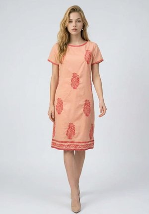 Mujer joven de pie con un vestido melocotón hasta la rodilla con patrones rojos y tacones altos beige sobre un fondo gris.