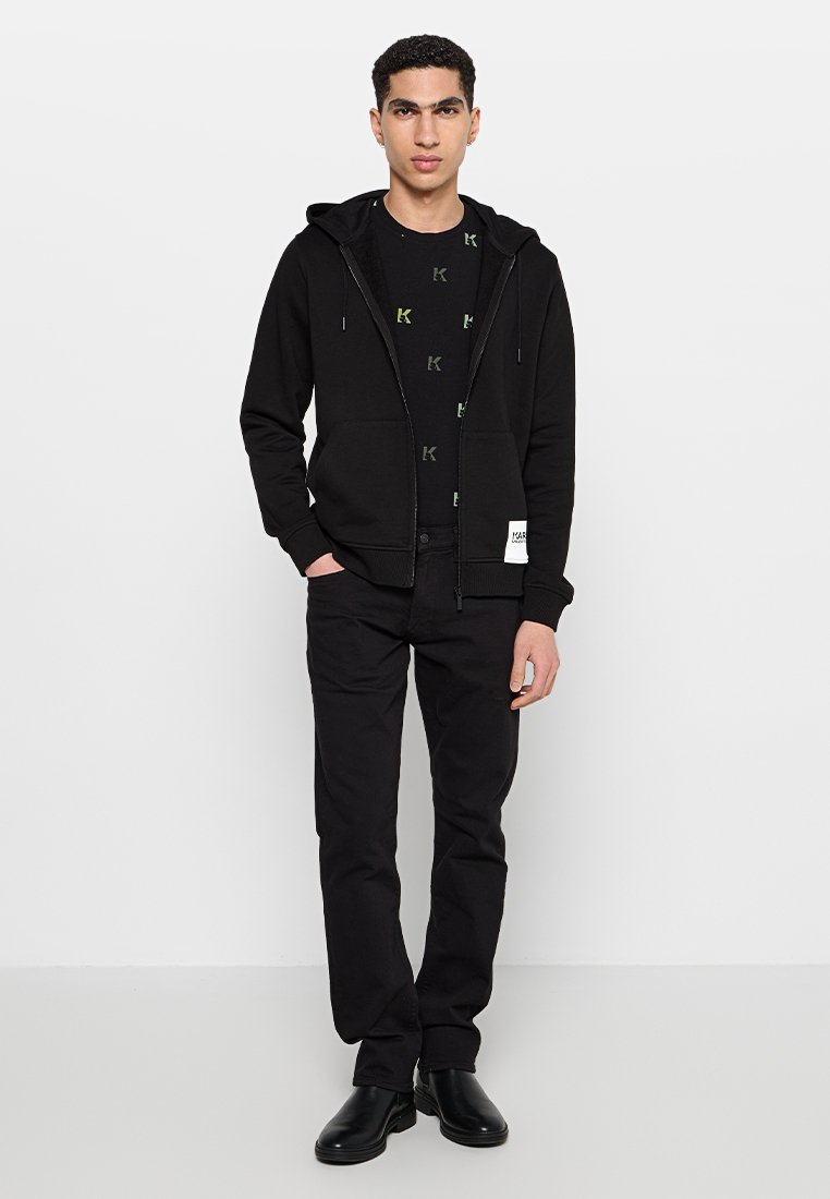 Sudadera negra con cremallera y bolsillo tipo canguro, llevada sobre una camiseta negra con patrones de logotipo en verde, combinada con pantalones y zapatos negros.