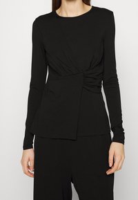 Czarna bluzka z długim rękawem z detalem w postaci skrętu w talii, wykonana z miękkiego materiału, z okrągłym dekoltem i gładką fakturą.