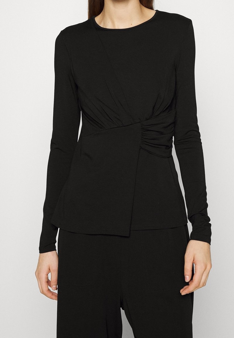 Czarna bluzka z długim rękawem z detalem w postaci skrętu w talii, wykonana z miękkiego materiału, z okrągłym dekoltem i gładką fakturą.