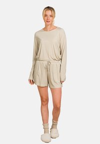 Helles beiges Loungewear-Set bestehend aus einem langärmeligen Oberteil mit einem dezenten Muster und Shorts mit Kordelzug. Kombiniert mit cremefarbenen, flauschigen Socken.