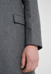 Gros plan sur la main d'une personne à côté de la manche et du rabat de poche d'un manteau en laine gris sur un fond neutre.