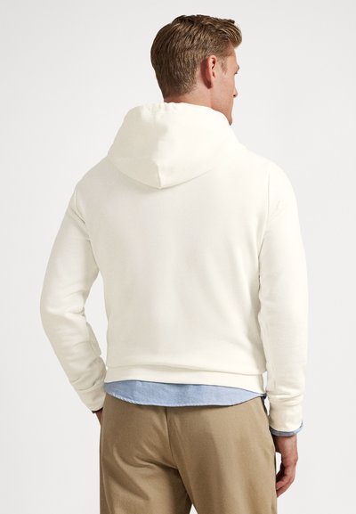 Φωτεινή κρεμ φούτερ με χαλαρή εφαρμογή, με κουκούλα με κορδόνι και μανσέτες με ρίγες. Φορεμένο πάνω από ένα ανοιχτό μπλε πουκάμισο, συνδυασμένο με μπεζ παντελόνια.