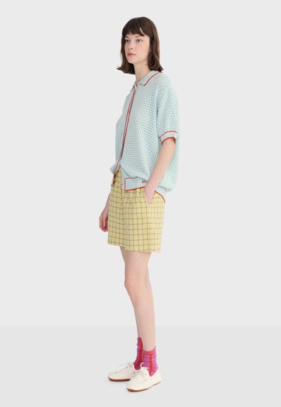 Femme debout portant un haut tricoté à manches courtes bleu clair avec bordure rouge, un short à carreaux jaunes, des chaussettes roses et des chaussures blanches, faisant face à gauche.