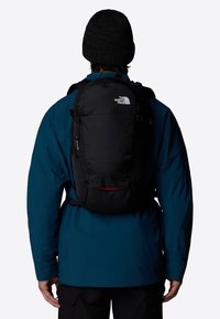 Mochila negra con textura suave, que presenta un logo prominente, cremalleras laterales y correas ajustables, llevada sobre una chaqueta azul.