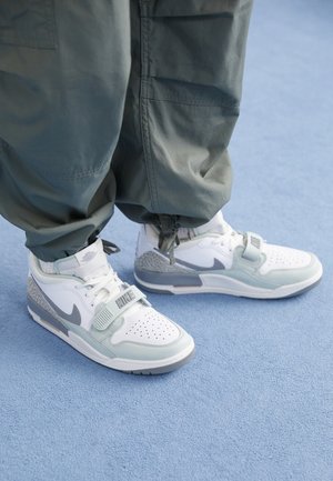 Baskets Nike blanches et grises avec lacets et sangle, portées avec un pantalon gris ample sur une surface texturée bleu clair.