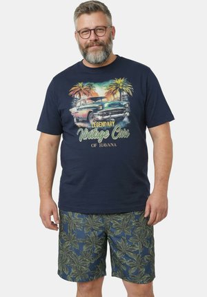 Baardige man met bril, een marineblauw T-shirt met een print van een vintage auto en palmbomen, en blauwe korte broek met groene palmbladeren patronen.