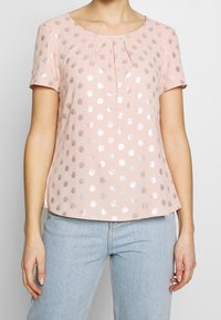 Top de manga corta y cuello redondo en suave tela rosa, con lunares plateados y un pliegue frontal para un detalle adicional. Combinado con un vaquero claro.