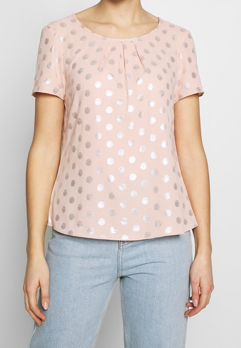 Top de manga corta y cuello redondo en suave tela rosa, con lunares plateados y un pliegue frontal para un detalle adicional. Combinado con un vaquero claro.