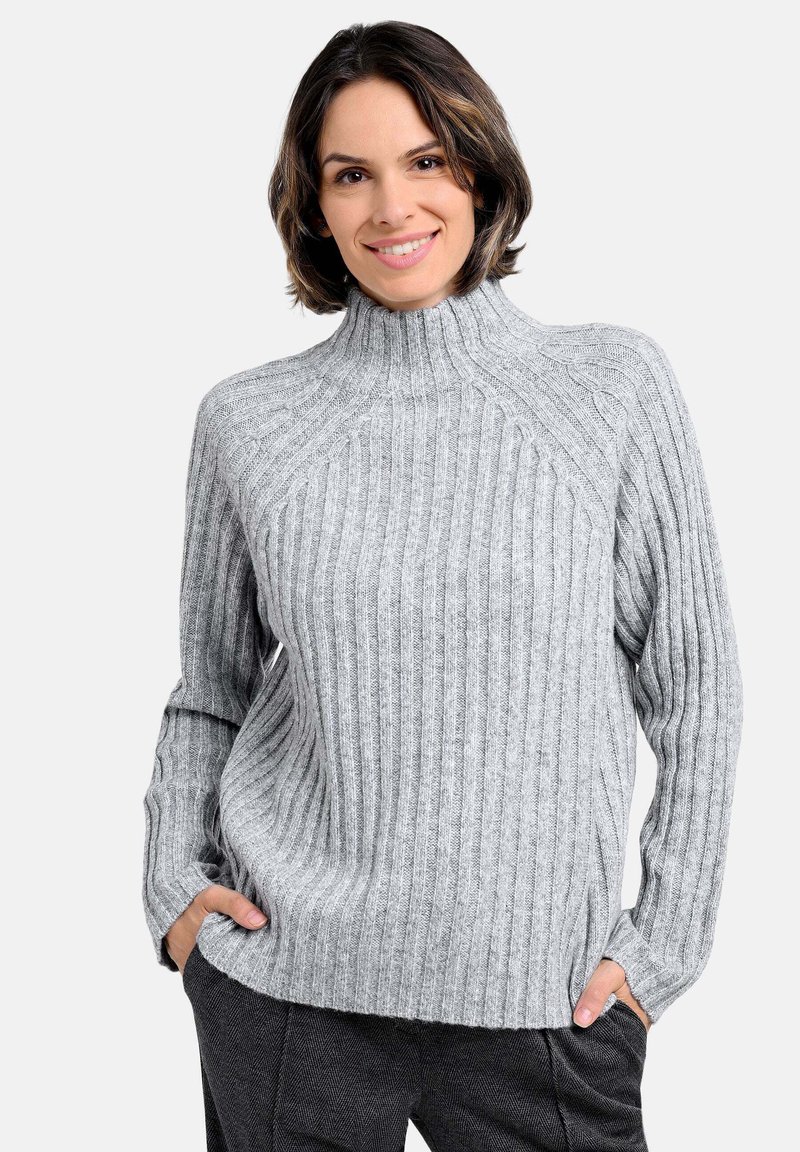 Grauer gerippter Pullover mit hohem Kragen, der ein strukturiertes Design und lange Ärmel aufweist. Das Modell posiert mit den Händen in den Taschen.