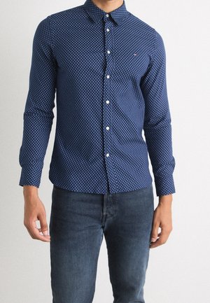 Hombre con una camisa entallada azul marino con estampado y botones, de manga larga, y vaqueros azul oscuro, sobre un fondo claro liso.