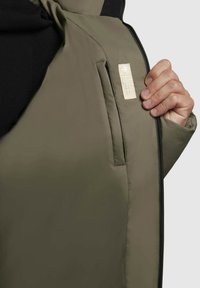 Olive zaļš jakas ar gludu tekstūru, ar sānu kabatu, kurai ir krēmveida etiķete un melna iekšējā odere. Redzams rāvējslēdzējs.