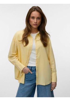 Frau mit langen braunen Haaren, die ein lockeres gelbes Hemd über einem weißen Top und blauen Jeans trägt, steht vor einem einfarbigen Hintergrund.