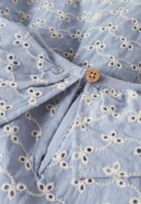 Vaaleansininen kangas, jossa valkoinen kukkakirjailu. Edessä nappikiinnitys pääntiellä ja koko pinnassa tekstuurikuvio.