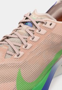 Beżowe sneakersy Nike z teksturowanego materiału, jasnoszare akcenty i niebieska podeszwa. Posiadają plecione sznurowadła oraz wyraźny detal z logo.