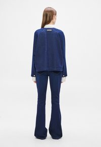 Μπλε τζιν μπουφάν με μακριά μανίκια, με λευκό γιακά και ταμπέλα, σε συνδυασμό με μπλε τζιν παντελόνια με καμπάνες και διακριτικές διακοσμητικές χρυσές κουμπιά.