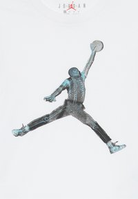 Biele bavlnené tričko s čiernymi a modrými grafikami basketbalistu v skoku, zobrazujúcim kostrový dizajn.