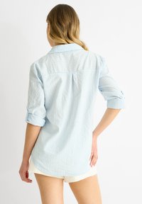 Camicia button-down a righe azzurro chiaro, con maniche lunghe arrotolate. Presenta una texture morbida e un orlo curvo, indossata con pantaloni corti bianchi.