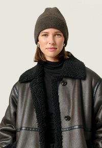 Braune Strickmütze, strukturiert, mit spitz zulaufendem Oberteil. Mit Lammfell gefütterte schwarze Lederjacke mit großem Kragen und glänzenden Knöpfen. Schwarzes Rollkragenoberteil darunter.